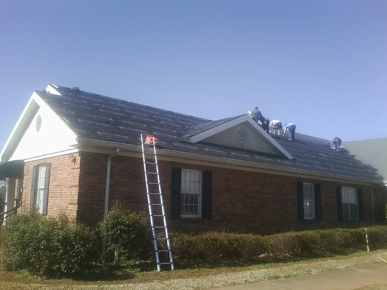 Crew tearing shingles off a brick ranch roof with a ladder set — Metal Roof Repair work by our crew in Avoca