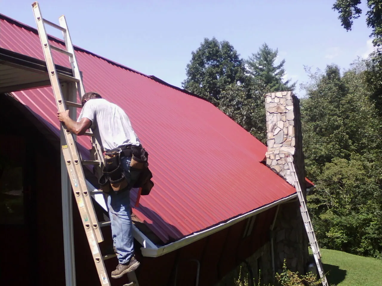 Roofer on a ladder beside a red metal roof and stone chimney — Soffit & Fascia Repair by Roofing Brothers in Avoca